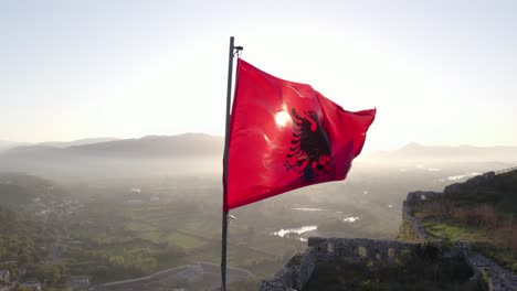 Orbit-around-Albanian-flag-waving-in-morning-sun-on-top-of-castle,-aerial