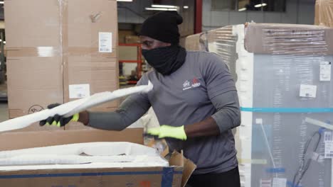 A-BIPOC-Ashley-Furniture-HomeStore-warehouse-merchandise-worker-uses-a-box-cutter-to-unpack-a-package-containing-cardboard-and-Styrofoam-on-a-workbench-inside-a-shipping-distribution-center