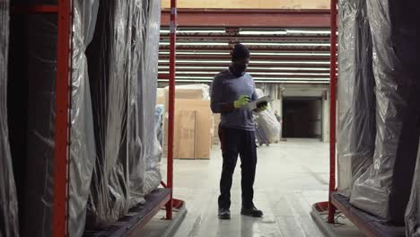 A-BIPOC-Ashley-Furniture-HomeStore-dufresne-warehouse-merchandise-worker-walks-between-two-isle-racks-inside-a-Canadian-distribution-center-using-his-note-board-to-check-stock-and-store-inventory