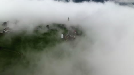 Aerial-view-through-thick-fog,-revealing-the-St-Nicholas-church-and-the-Renon-town,-in-Italy