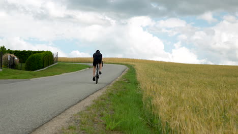 Professional-road-cyclist-man-cycling-uphill-on-a-winding-country-road-by-crop-field-low-angle-rear-view