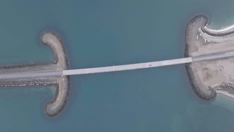 Top-Down-Aerial-View-of-Car-Passing-Bridge-Between-Barrier-and-Breakwater-on-Glacial-Water-in-Landscape-of-Iceland
