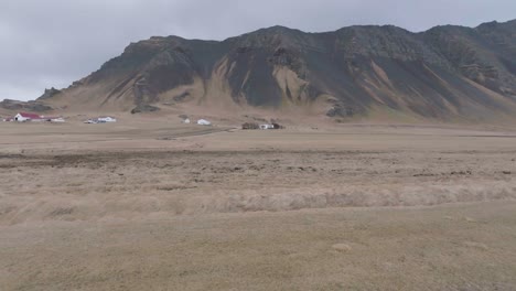 Flug-über-Gras-Und-Feuchtem-Land-In-Der-Isländischen-Landschaft,-Dorfhäusern-Und-Hügeln-Im-Hintergrund,-Drohnenaufnahme