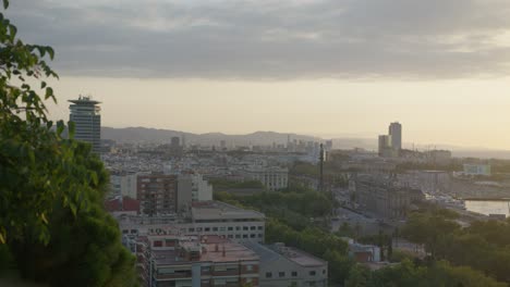 Hochauflösende-Ansicht-Durch-Malerische-Bäume,-2.-Winkel,-Skyline-Der-Stadt-Barcelona,-Spanien,-Mit-Goldenem-Sonnenaufgang-In-6K,-Während-Vögel-Fliegen