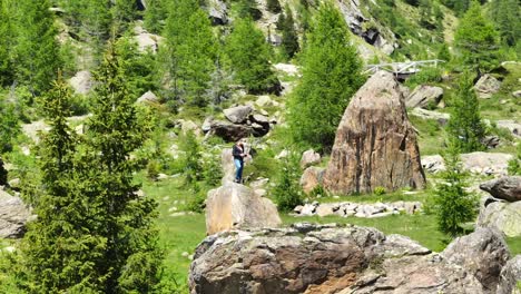 Man-standing-on-rock-with-drone-remote-controller-surrounded-by-nature-of-Val-Masino-in-Valtellina,-Italy