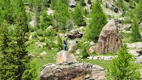 Man-standing-on-rock-with-drone-remote-controller-surrounded-by-nature-of-Val-Masino-in-Valtellina,-Italy
