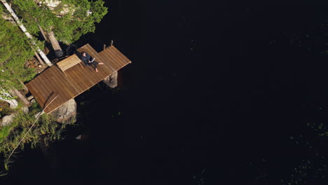 Aerial-descending-view-of-a-woman-sitting-on-a-deck-reading-a-book-on-the-water