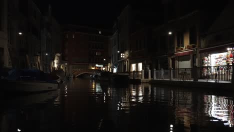 Beautiful-Venice-Canal-Waters-At-Night-With-Light-Of-Buildings-And-Shops-Reflected-On-Waters-Surface-With-Silhouette-Of-People-Walking-Past