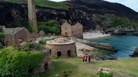 Aerial-view-panning-across-Porth-Wen-abandoned-brickwork-factory-with-tourists-talking-outside-overgrown-brick-furnace