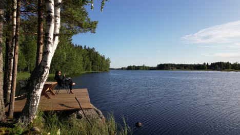 Mujer-Sentada-Con-Una-Copa-De-Vino-Y-Un-Libro-En-La-Cubierta-Junto-Al-Mar,-Vacaciones-En-Finlandia