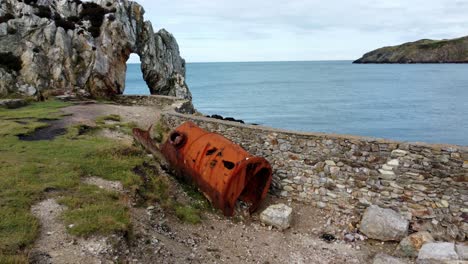 Aerial-view-circling-red-rusted-industrial-tank-to-reveal-rock-archway-on-the-Welsh-coastline