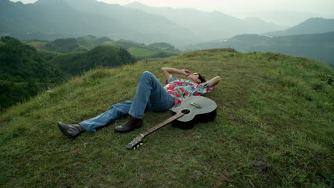 Male-Musician-with-Acoustic-Guitar-Lays-Down-at-Green-Mountain-Hills-Landscape-watching-the-Sky,-wearing-Vintage-Colorful-Clothes