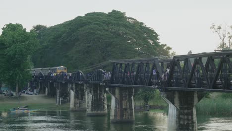 Un-Tren-Que-Cruza-El-Puente-Del-Río-Kwai-Por-La-Noche,-Mientras-Los-Turistas-Observan-De-Pie-En-El-Famoso-Puente-De-La-Segunda-Guerra-Mundial-En-Kanchanaburi,-Tailandia
