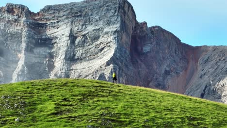 Tight-162mm-drone-shot-of-a-female-hiker-walking-on-a-hill-with-a-giant-vertical-rock-face-of-a-mountain-behind-her-in-the-Dolomites