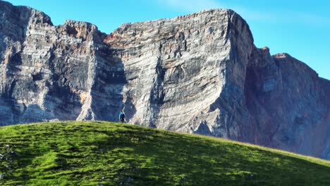 Tight-162mm-drone-shot-of-a-male-hiker-walking-on-a-hill-with-a-dramatic-vertical-rock-face-in-the-background-in-the-Dolomite-mountains-of-Italy