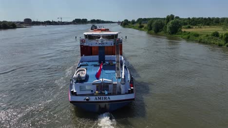 Ein-Amira-Boot-Mit-Containern,-Das-Sich-Auf-Einem-Fluss-Mit-Bäumen-An-Den-Ufern-Bewegt