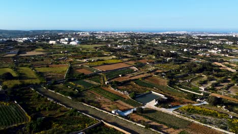 Drone-view-of-Maltese-city-in-the-distance-with-the-countryside-in-the-foreground,-ocean-visible-on-horizon