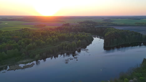 Drone-view-flying-over-a-spectacular-countryside-of-a-forest,-lake,-canals,-open-lands-and-a-majestic-sunset