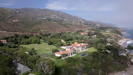Aerial-view-through-clouds,-revealing-a-mansion-on-the-coast-of-Malibu,-CA,-USA