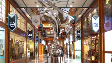 Static-shot-at-iconic-Brisbane-arcade,-shopping-galleria-in-heritage-listed-building-in-Queen-Street-Mall,-capturing-shoppers-window-shopping-and-Mirage-sculpture-by-Gidon-Graetz-as-the-centrepiece