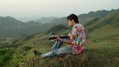 Asiatischer-Männlicher-Gitarrist-Spielt-In-Der-Malerischen-Landschaft-Der-Grünen-Hügel,-Musiker-In-Vintage-Kleidung,-Hawaiihemd-Und-Sonnenbrille