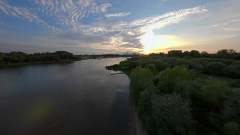 FPV-fly-over-the-river-bank-of-the-shore-of-Vistula-river-chasing-the-sunset-with-the-skyline-of-Warsaw,-Poland-in-the-background