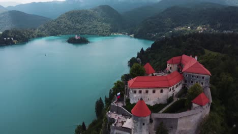 Aerial-tilt-up-shot-of-famous-castle-at-bled-lake-with-waving-Slovenian-flag-and-church-on-island-in-Background