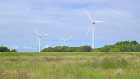 Wind-turbines-rotating-with-tracking-across-grassy-meadow-ending-with-rubbish-pile-on-cloudy-summer-day