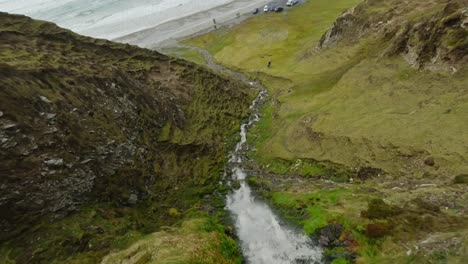 Drone-diving-down-a-small-waterfall-on-green-cliffs-towards-Keel-Beach-in-Ireland