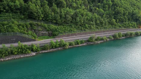 Drone-arc-shot-of-scenic-road-amidst-beautiful-turquoise-lake-and-lush-green-forest