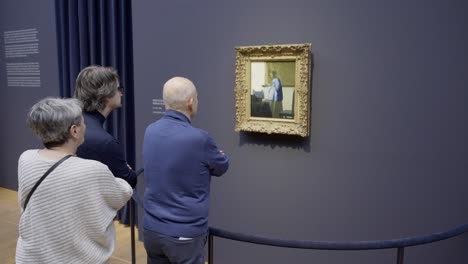 Los-Visitantes-Cautivados-Se-Maravillan-Con-&#39;la-Lechera&#39;-De-Vermeer-En-La-Exhibición-Vermeer-Del-Rijksmuseum,-Elegantemente-Enmarcada-Contra-Un-Fondo-Azul-Sereno