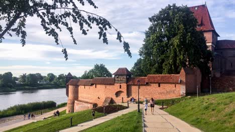 Nogat-river-waterfront-in-Malbork,-Poland-at-beautiful-sunset-with-a-castle,-the-red-brick-walls-and-tourist-people