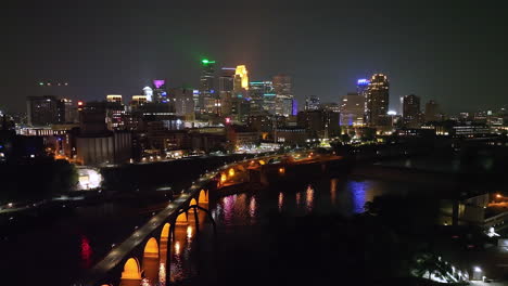 Minneapolis-illuminated-city-skyline-view,-night-aerial-over-Mississippi-river