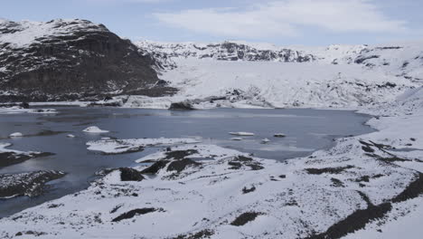 Aufsteigende-Luftaufnahme-Eines-Zugefrorenen-Isländischen-Sees-Am-Fuße-Eines-Riesigen-Gebirgstals