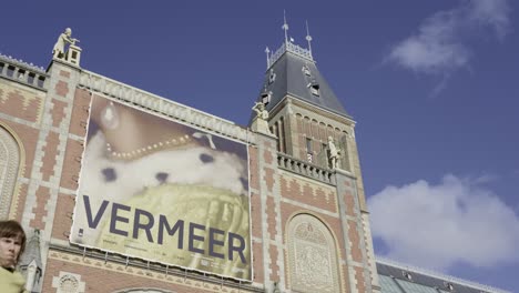 Esplendor-Arquitectónico:-Primer-Plano-Del-Edificio-Del-Rijksmuseum-En-Medio-De-La-Dinámica-Vida-De-La-Ciudad,-Envuelto-Por-Nubes-Y-Adornado-Con-Una-Impresión-De-Exposición-De-Vermeer