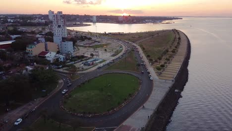 Aerial-view-of-city,-waterfront,-and-cars-driving-at-sunset-in-beautiful-Posadas,-Misiones,-Argentina