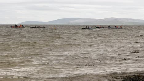Currach-boats-race-through-ocean-with-county-clare-behind,-static