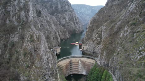 Drone-view-of-dam-and-building-on-Treska-River-passing-through-Matka-Canyon,-athletes-canoeing-on-the-river