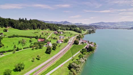 Luftflug-Entlang-Der-Küste-Des-Obersees-In-Idyllischer-Landschaft-Mit-Kleinem-Dorf-Und-Kirche-In-Der-Schweiz-An-Sonnigen-Tagen