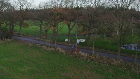 Drone-shot-of-man-cycling-on-small-road-with-trees-on-the-side-during-autumn,-aerial