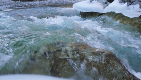 Emerald-river-water-at-the-winter-time-flowing-through-rocks-under-snow,-slow-motion