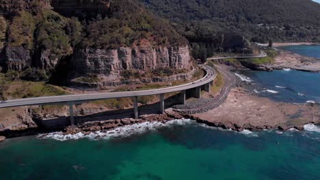 Aerial-view-of-cars-driving-on-Sea-Cliff-Bridge,-sunny-day,-grand-pacific-drive,-New-South-Wales,-Australia---orbit-drone-shot