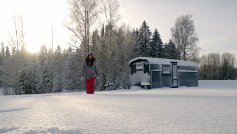 Woman-in-grey-jacket-red-pants-walks-across-barren-snowy-landscape-in-front-of-forest