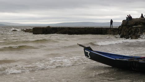 Currach-boat-bow-sits-above-water-as-spectators-roam-ladies-beach,-static