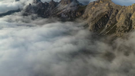Drone-shot-over-low-hanging-clouds,-tilting-toward-rocky-peaks-of-Dolomites,-Italy