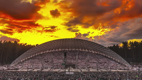 Timelapse-shot-of-performance-underway-surrounded-by-huge-crowd-during-Latvian-Song-and-Dance-Festival-in-an-outdoor-stadium-in-Riga,-Latvia-with-yellow-sky-in-the-background