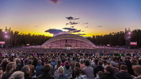 Timelapse-shot-of-huge-crowd-enjoying-the-Latvian-Song-and-Dance-Festival-in-an-outdoor-stadium-in-Riga,-Latvia-with-yellow-evening-sky-in-the-background