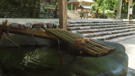 Agua-Que-Sale-De-Una-Tubería-De-Bambú-Hacia-Una-Fuente-Con-Un-Par-De-Cucharones-De-Madera-Descansando-Cerca-Del-Santuario-En-Shirakawago