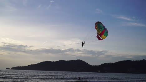 Tracking-shot-of-tourist-parasailing-over-coast-and-water-at-Patong-Beach-during-golden-sunset-in-Thailand