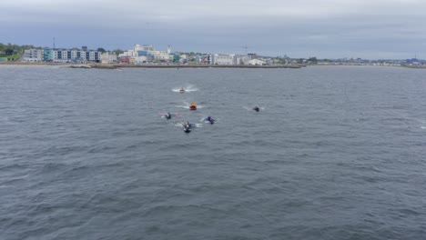 Aerial-frontal-tracking-view-of-boats-bobbing-paddling-currachs-across-open-ocean,-galway-in-background